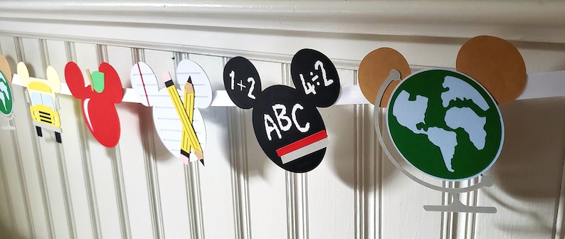 May include: A white banner with Mickey Mouse ears features various school-themed images, including a school bus, apple, pencils, a chalkboard with the text "1+2" and "4:2", a chalkboard with the text "ABC", and a globe.