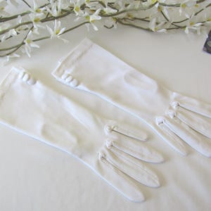 May include: A pair of elegant white gloves, likely for formal occasions, are laid out on a white surface. The gloves have a smooth texture, with button details at the wrist and finger seams. The gloves are long, extending past the wrist.
