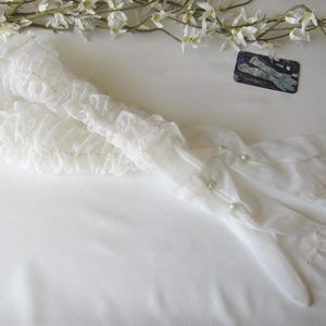 Peut inclure: Une paire d'élégants gants blancs transparents avec de délicates volants et des perles. Les gants sont longs, s'étendant au-delà du poignet, et sont faits d'un tissu léger et fluide. Une branche de fleurs blanches est en arrière-plan.