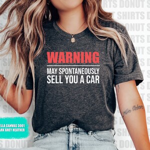 May include: A dark grey heather Bella Canvas 3001 t-shirt with the text "WARNING MAY SPONTANEOUSLY SELL YOU A CAR" in red and white lettering.