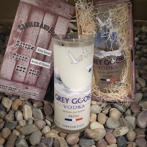 May include: A white candle in a clear glass bottle shaped like a Grey Goose Vodka bottle. The candle has a white label with the text "Grey Goose Vodka" and "Distilled and Bottled France Imported". The candle is sitting on a bed of small, colorful rocks.