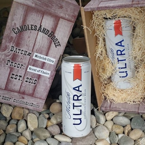 May include: A wooden crate with the words "Candles and Booze" printed on the side. The crate contains two cans of Michelob Ultra beer. The cans are white with blue lettering and a red stripe. The text on the can reads "Michelob Ultra" and "Superior Light Beer".