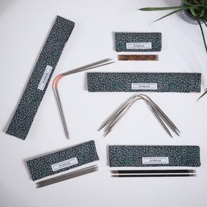 Puede incluir: Una colección de agujas de tejer y accesorios, que incluyen agujas rectas, circulares y de doble punta. Los artículos se muestran con estuches con estampado de leopardo verde azulado. Cada estuche tiene una etiqueta que dice "AnMiwe."