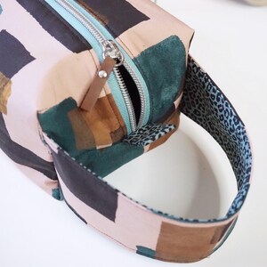May include: A rectangular toiletry bag featuring a light pink base with abstract black, dark green, and brown brushstroke patterns. It includes a light blue zipper with a brown leather pull tab and a handle lined with a blue and black leopard print.