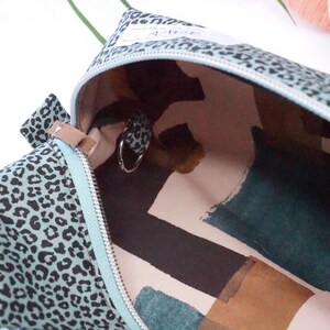 May include: A zippered pouch featuring a light blue exterior with a black leopard print pattern. The interior is lined with a cream fabric displaying abstract shapes in dark green, black, and brown. A small metal ring is attached to the inner side.