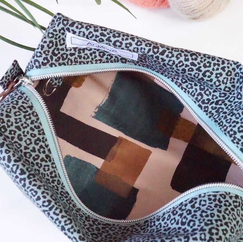 May include: A zippered pouch featuring a teal and black leopard print exterior with a white label reading AnMiwe. The interior is lined with a fabric displaying a geometric pattern of dark teal, brown, and cream-colored rectangular shapes.