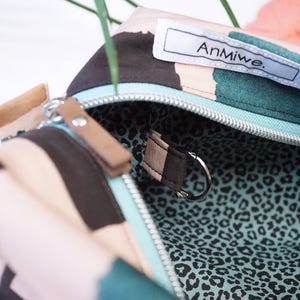 May include: A zippered pouch with a multi-colored abstract pattern in shades of pink, dark brown, and teal. The interior is lined with a teal and black leopard print fabric and features a small metal D-ring. A white label reads AnMiwe.