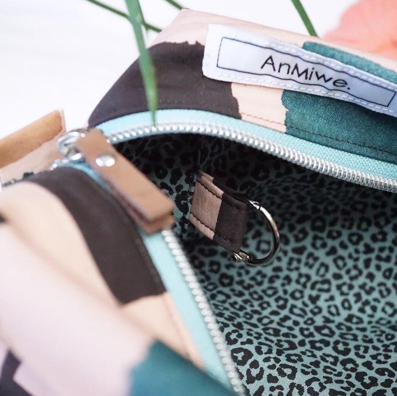 May include: A zippered pouch with a multi-colored abstract pattern in shades of pink, dark brown, and teal. The interior is lined with a teal and black leopard print fabric and features a small metal D-ring. A white label reads AnMiwe.