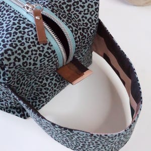 May include: A rectangular toiletry bag featuring a light blue fabric with a black leopard print pattern. The bag includes a silver metal zipper with a brown leather pull tab and a matching fabric handle attached to the side with a brown leather accent.