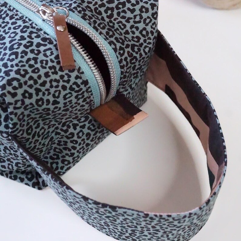 May include: A rectangular toiletry bag featuring a light blue fabric with a black leopard print pattern. The bag includes a silver metal zipper with a brown leather pull tab and a matching fabric handle attached to the side with a brown leather accent.