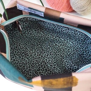 May include: A zippered pouch featuring an exterior with abstract black, teal, and gold brushstroke patterns on a light background. The interior is lined with a light blue fabric printed with a black leopard spot pattern. A white label reads Anmiwe.