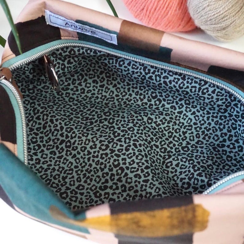 May include: A zippered pouch featuring an exterior with abstract black, teal, and gold brushstroke patterns on a light background. The interior is lined with a light blue fabric printed with a black leopard spot pattern. A white label reads Anmiwe.