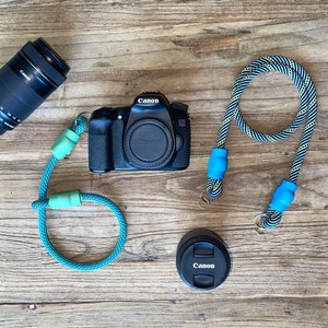 May include: A black Canon EOS 70D camera body with a lens cap, a separate Canon lens, a detached lens cap, and two camera straps. One strap is solid teal with light green connectors, and the other is a black and white patterned rope with blue connectors.
