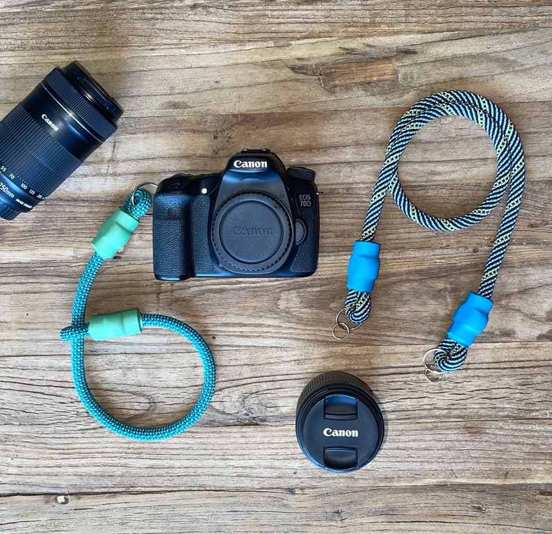 May include: A black Canon EOS 70D camera body with a lens cap, a separate Canon lens, a detached lens cap, and two camera straps. One strap is solid teal with light green connectors, and the other is a black and white patterned rope with blue connectors.
