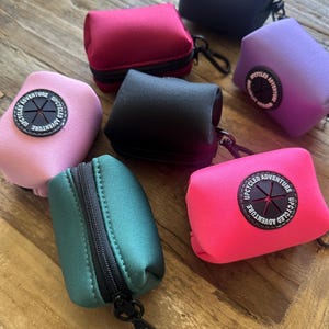May include: Assortment of seven small zippered pouches in various colours, including pink, green, black, purple, red, and blue. Each pouch has a black zipper and a circular logo with the text "Upcycled Adventure" and a starburst design.