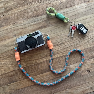 May include: A silver and black Petri camera attached to a blue and orange rope strap with peach-coloured connectors. A separate lime green and black rope keychain features a red paw print charm, two metal keys, and a black electronic car key fob.