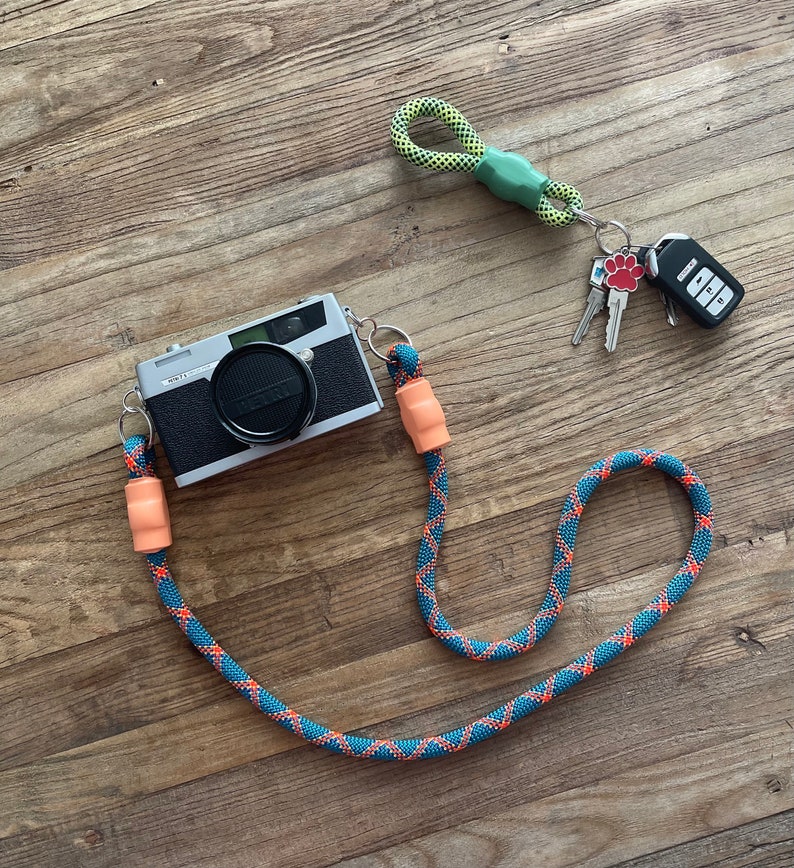 May include: A silver and black Petri camera attached to a blue and orange rope strap with peach-coloured connectors. A separate lime green and black rope keychain features a red paw print charm, two metal keys, and a black electronic car key fob.