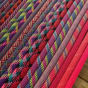 May include: A collection of braided ropes arranged in parallel lines, featuring a vibrant spectrum of colours including neon pink, purple, blue, yellow, and orange. Each rope displays a distinct woven texture and intricate geometric patterns.
