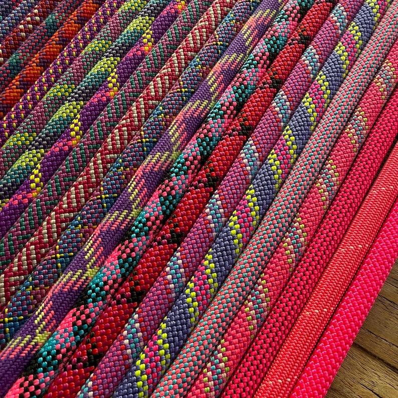 May include: A collection of braided ropes arranged in parallel lines, featuring a vibrant spectrum of colours including neon pink, purple, blue, yellow, and orange. Each rope displays a distinct woven texture and intricate geometric patterns.