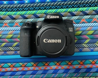 May include: Black Canon EOS 70D digital camera with lens cap, set against a backdrop of colourful, patterned ropes. The Canon logo is visible on the camera body and lens cap. The ropes are in shades of blue, green, and red.