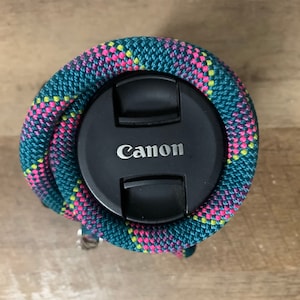 May include: A black Canon camera lens cap surrounded by a thick, braided rope strap featuring a vibrant pattern of teal, hot pink, and bright yellow woven threads. The circular cap is centred within the coiled, textured rope accessory.