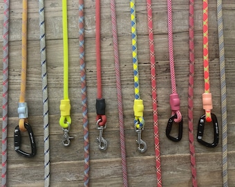 May include: A collection of vertical climbing ropes in various colours and patterns, including solid neon yellow, red, and multi-coloured braided designs. Each rope features a metal carabiner or a swivel snap hook attached with a coloured sleeve.