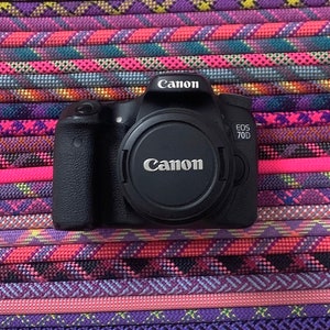 May include: A black Canon EOS 70D digital camera body with a lens cap attached. The camera features the Canon logo on the top prism housing and the lens cap, with the model name EOS 70D printed in white on the right side of the camera body.