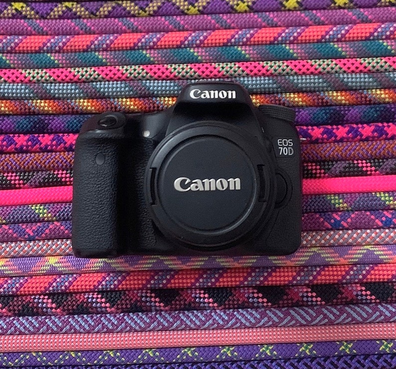 May include: A black Canon EOS 70D digital camera body with a lens cap attached. The camera features the Canon logo on the top prism housing and the lens cap, with the model name EOS 70D printed in white on the right side of the camera body.