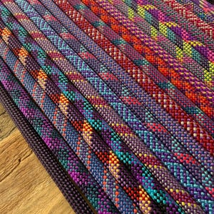 May include: Multiple strands of braided rope arranged in parallel rows. Each rope features a complex, multi-coloured woven pattern incorporating vibrant shades of purple, magenta, orange, yellow, and teal, creating a textured, repeating geometric design.