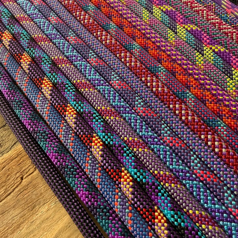 May include: Multiple strands of braided rope arranged in parallel rows. Each rope features a complex, multi-coloured woven pattern incorporating vibrant shades of purple, magenta, orange, yellow, and teal, creating a textured, repeating geometric design.