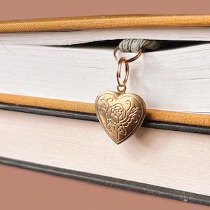 Könnte beinhalten: Ein goldener herzförmiger Lesezeichen mit floralen Details hängt zwischen den Seiten eines Buches. Das Buch hat einen senfgelben Einband und weiße Seiten. Darunter liegt ein zweites Buch mit schwarzem Einband.