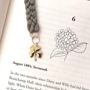 Op de afbeelding: Een gevlochten grijze bladwijzer met een goudkleurige klaverbedel, in een open boek. De pagina van het boek toont tekst en een bloementekening. Een decoratief accessoire voor lezers.