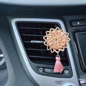 Puede incluir: Un ambientador de coche tejido en forma de estrella con un borla rosa colgando. El ambientador está sujeto a la ventilación del coche.