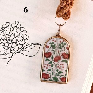 Könnte beinhalten: Ein goldfarbener Lesezeichen mit einem rechteckigen Anhänger mit einem Blumenmuster in Rot und Grün auf weißem Hintergrund. Das Lesezeichen hat eine geflochtene braune Kordel und befindet sich in einem offenen Buch. Die Zahl "6" ist auf der Seite zu sehen.