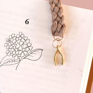 Könnte beinhalten: Geflochtener beigefarbener Makramee-Lesezeichen mit einem goldfarbenen Glücksbringer-Anhänger. Das Lesezeichen befindet sich in einem Buch mit einer Blumenillustration und der Zahl 6. Die Seiten des Buches sind sichtbar.
