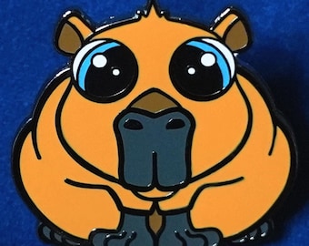 May include: Orange and black enamel pin featuring a cartoon capybara with large, round eyes. The capybara is sitting with its paws tucked in front of it. The pin is part of a collection of animal pins that support animal welfare.