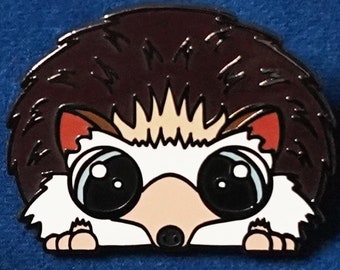 Puede incluir: Un pin de esmalte marrón y blanco de un erizo con ojos grandes que asoman por detrás de sus espinas. El pin forma parte de una colección de pines de animales que apoyan el bienestar animal.