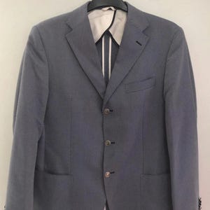 May include: A light blue blazer with a subtle, textured pattern. The jacket features a notched lapel, three-button closure, and two front pockets. The interior lining is visible, with a white and striped design. The blazer is hanging on a black hanger.