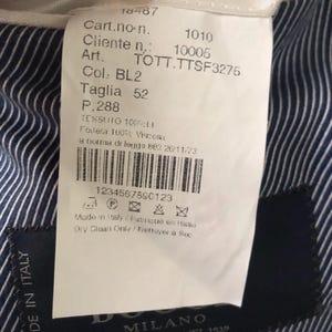 May include: A white tag with black text, attached to a blue and white striped fabric. The tag includes the text "MADE IN ITALY", "Taglia 52", and a barcode. The brand name "DO" and "MILANO" are also visible.
