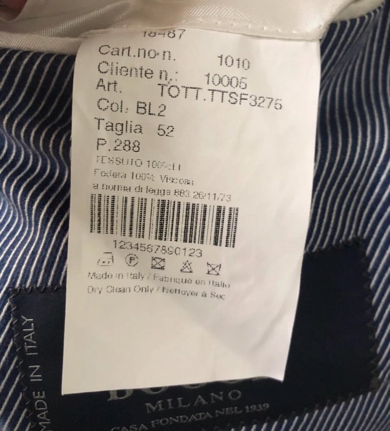 May include: A white tag with black text, attached to a blue and white striped fabric. The tag includes the text "MADE IN ITALY", "Taglia 52", and a barcode. The brand name "DO" and "MILANO" are also visible.