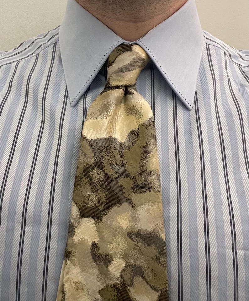 Puede incluir: Una corbata con un patr&oacute;n abstracto y moteado en tonos beige, verde oliva y marr&oacute;n oscuro, combinada con una camisa de vestir azul claro con rayas verticales blancas y azul oscuro y un cuello puntiagudo y estructurado.