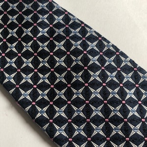 Pu&ograve; includere: Una cravatta con una base blu navy scuro e un motivo geometrico ripetitivo di forme a diamante bianche e piccoli quadrati rosa a ogni intersezione. Il tessuto presenta una trama testurizzata, creando un aspetto strutturato su tutta la superficie.
