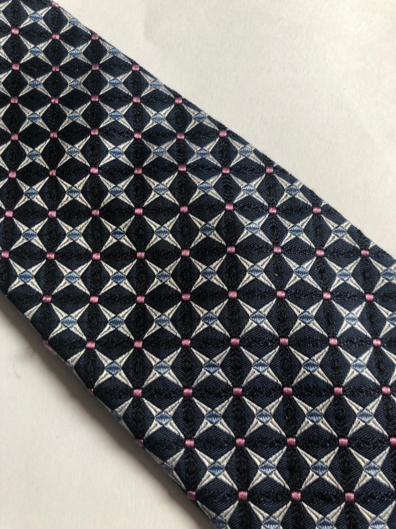 Pu&ograve; includere: Una cravatta con una base blu navy scuro e un motivo geometrico ripetitivo di forme a diamante bianche e piccoli quadrati rosa a ogni intersezione. Il tessuto presenta una trama testurizzata, creando un aspetto strutturato su tutta la superficie.
