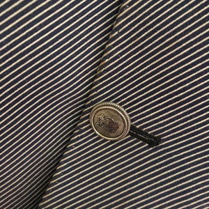 May include: Close-up of a navy blue and white striped blazer. The blazer features a decorative silver button with an embossed design and a black button loop. The fabric has a diagonal stripe pattern.