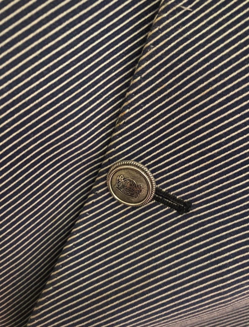 May include: Close-up of a navy blue and white striped blazer. The blazer features a decorative silver button with an embossed design and a black button loop. The fabric has a diagonal stripe pattern.