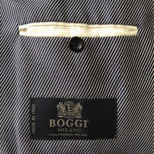 May include: Close-up of the interior of a navy blue and white striped blazer. A white pocket lining and a black button are visible. The BOGGI MILANO label is sewn into the lining, with text that reads "MADE IN ITALY" and "CASA FONDATA NEL 1939."