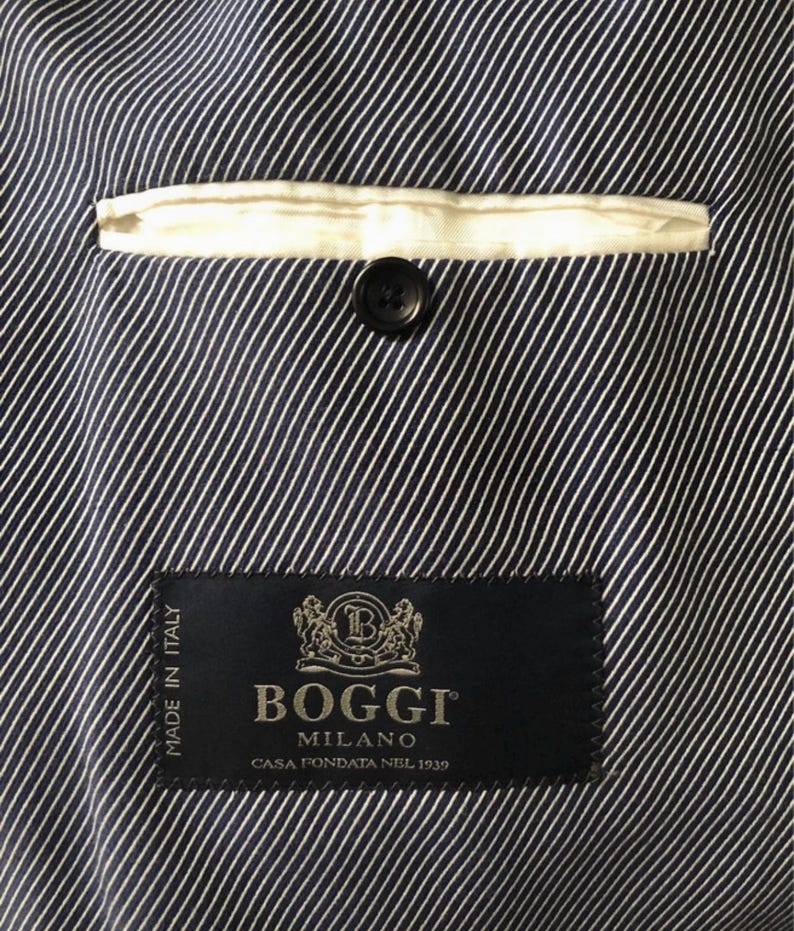 May include: Close-up of the interior of a navy blue and white striped blazer. A white pocket lining and a black button are visible. The BOGGI MILANO label is sewn into the lining, with text that reads "MADE IN ITALY" and "CASA FONDATA NEL 1939."