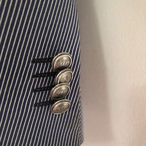 May include: Close-up of a navy blue and white striped blazer. The blazer features four silver-colored buttons with a detailed design. The buttons are on the sleeve of the blazer.