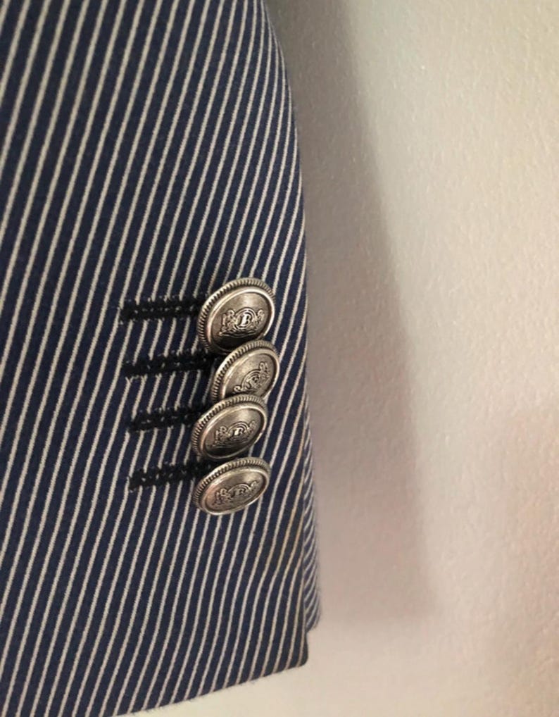 May include: Close-up of a navy blue and white striped blazer. The blazer features four silver-colored buttons with a detailed design. The buttons are on the sleeve of the blazer.