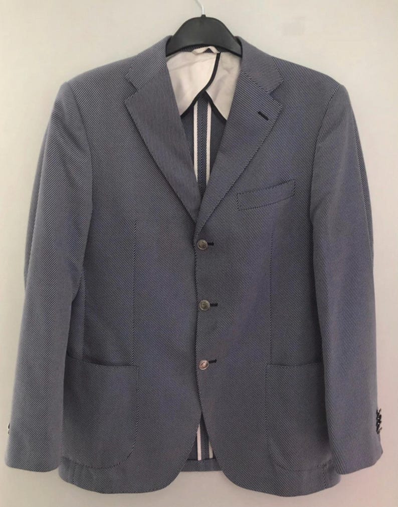 May include: A light blue blazer with a subtle, textured pattern. The jacket features a notched lapel, three-button closure, and two front pockets. The interior lining is visible, with a white and striped design. The blazer is hanging on a black hanger.